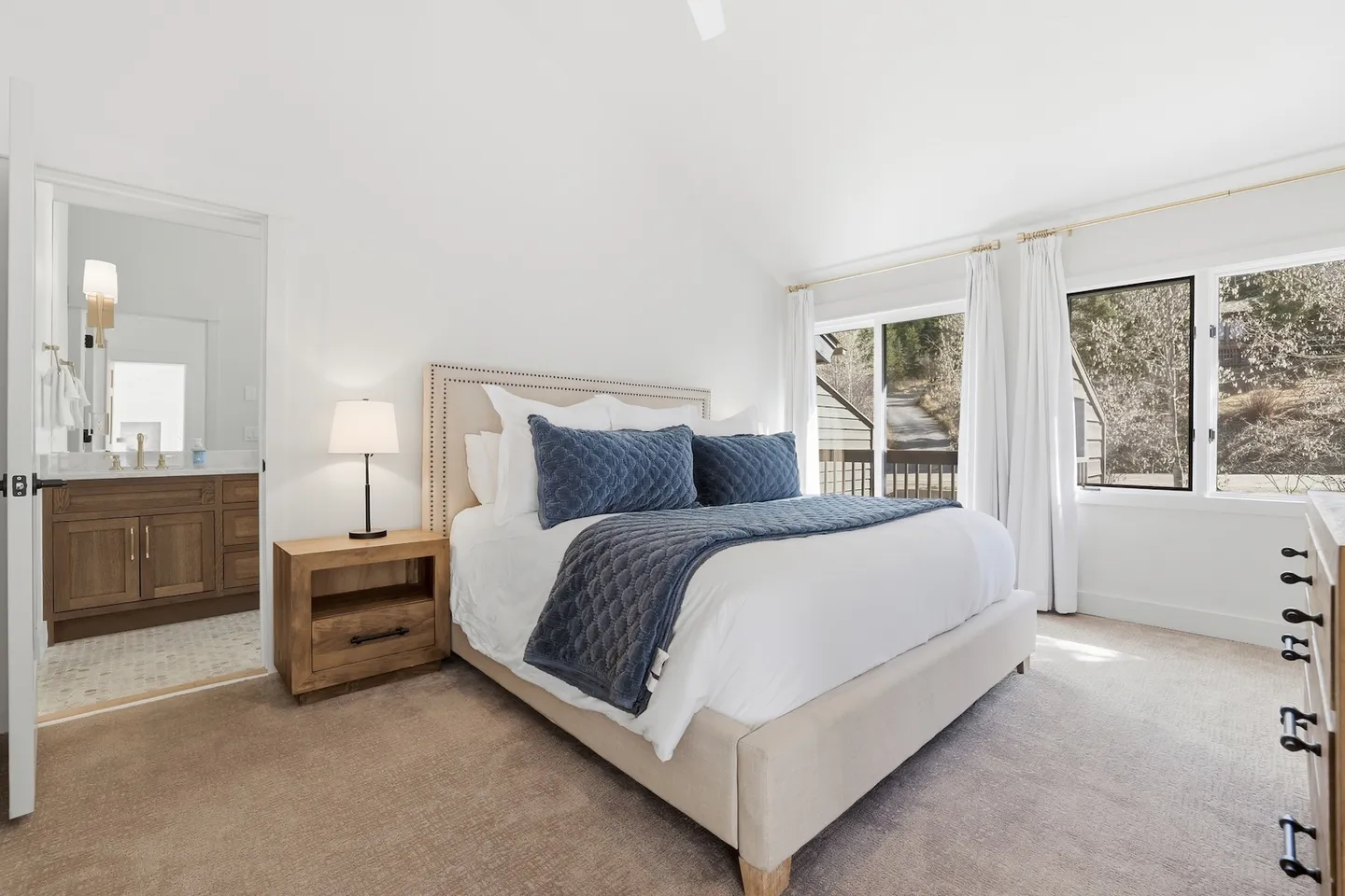 Bedroom with an upholstered bed, bedside nightstand with lamp, carpeted floor, sliding glass door, and an open doorway to an ensuite bathroom.