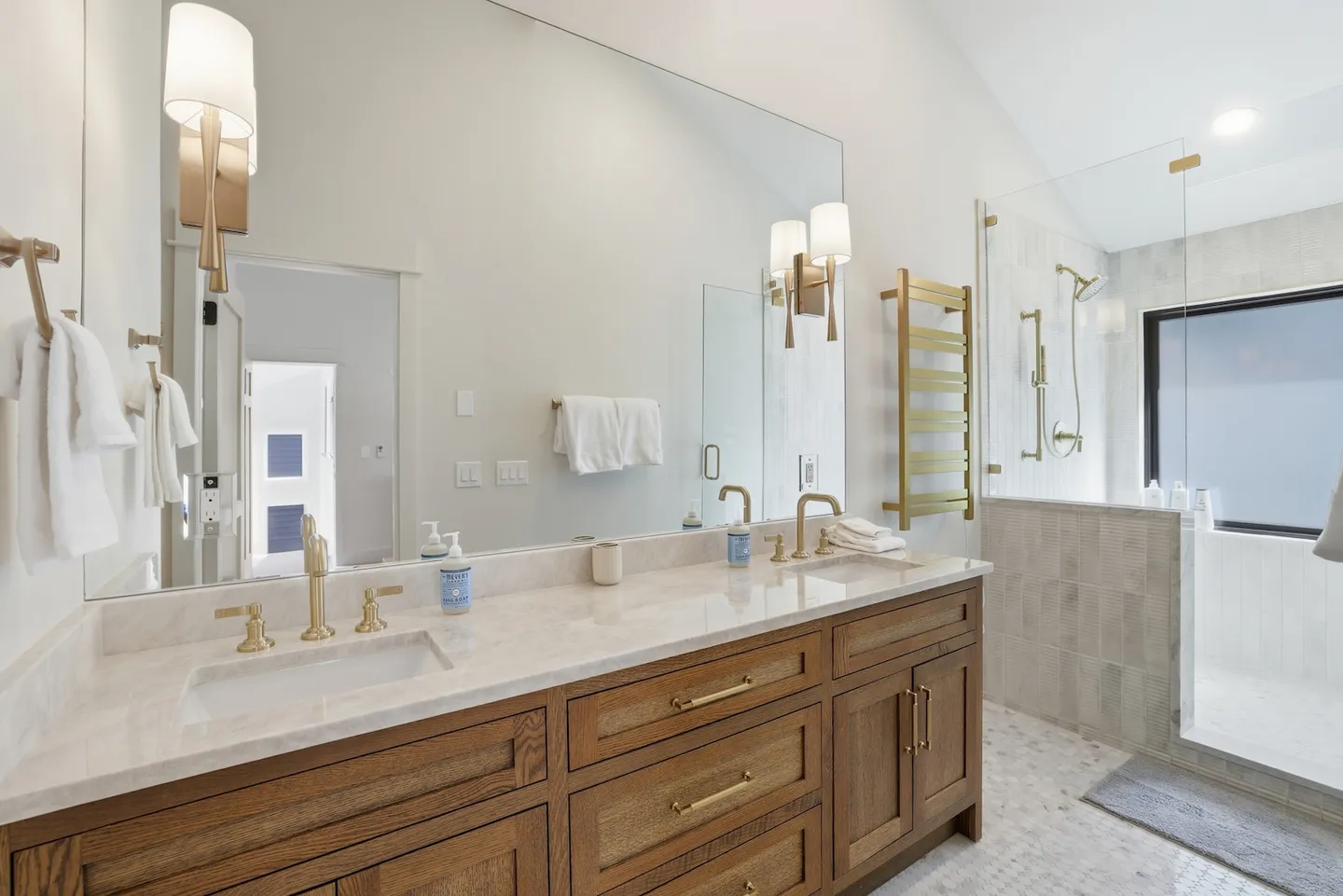 Bathroom with a double-sink wood vanity, brass fixtures, a large wall mirror, and a walk-in glass shower.