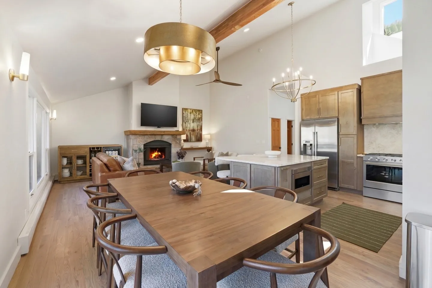 Open-concept dining, kitchen, and living area with wood dining table, pendant lighting, stone fireplace, and stainless steel appliances.