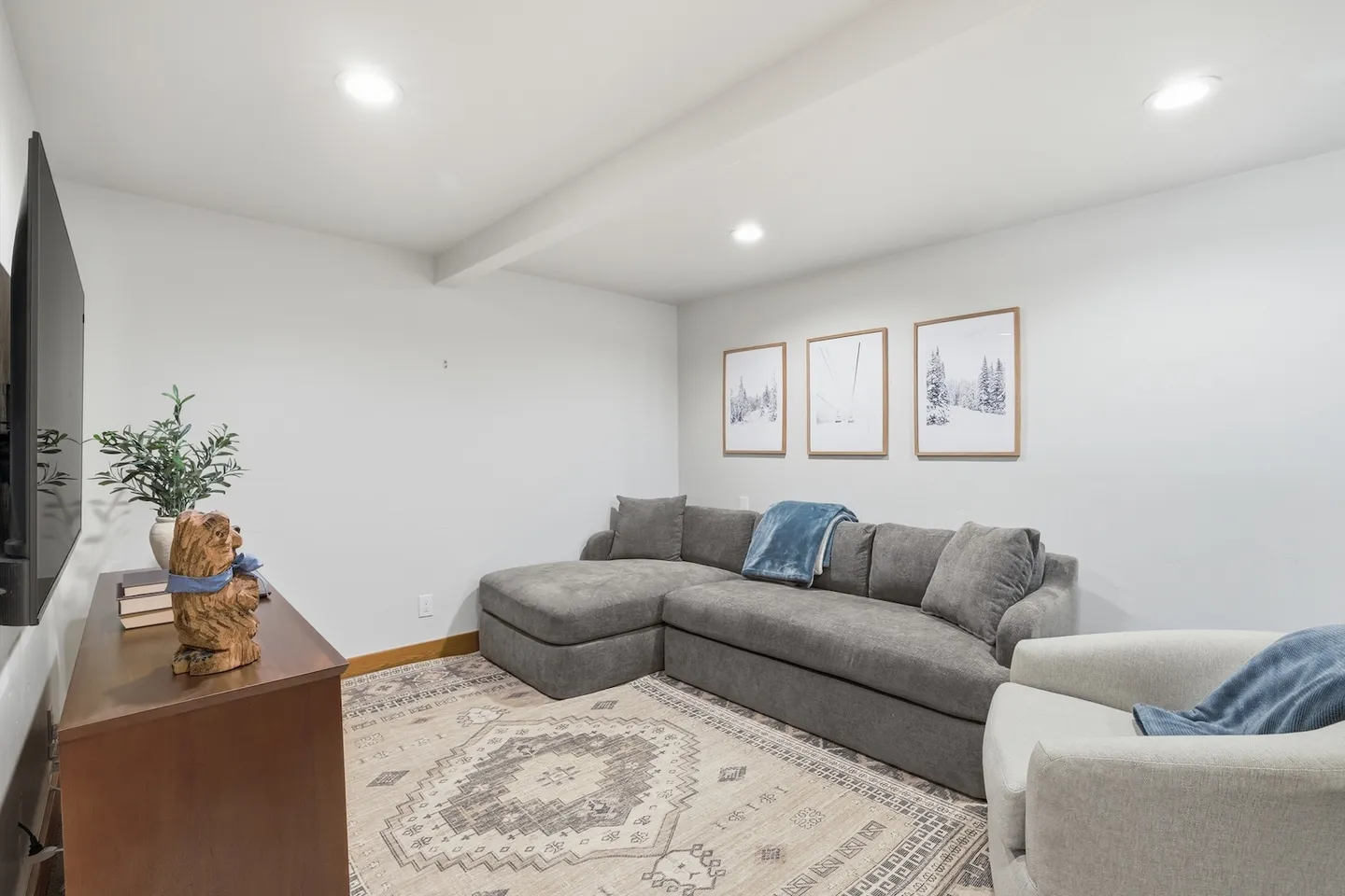 A living room area featuring a gray sectional sofa, patterned area rug, armchair, and media console below a wall-mounted TV.