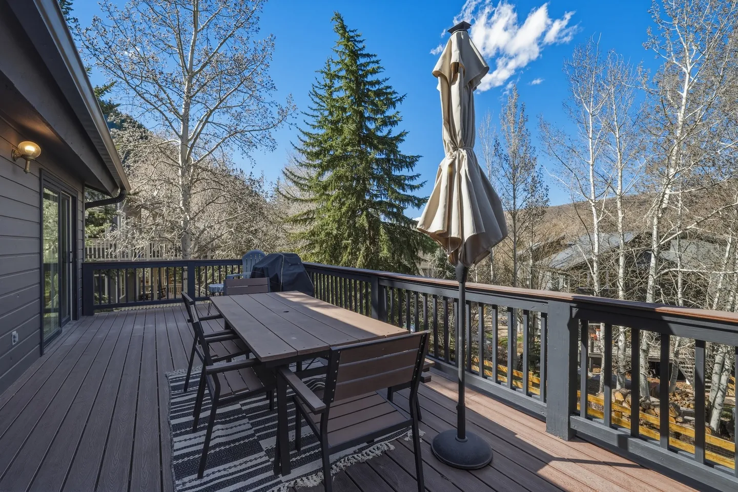 Outdoor deck with an outdoor dining table, chairs, a closed patio umbrella, and surrounding pine and aspen trees under a blue sky.