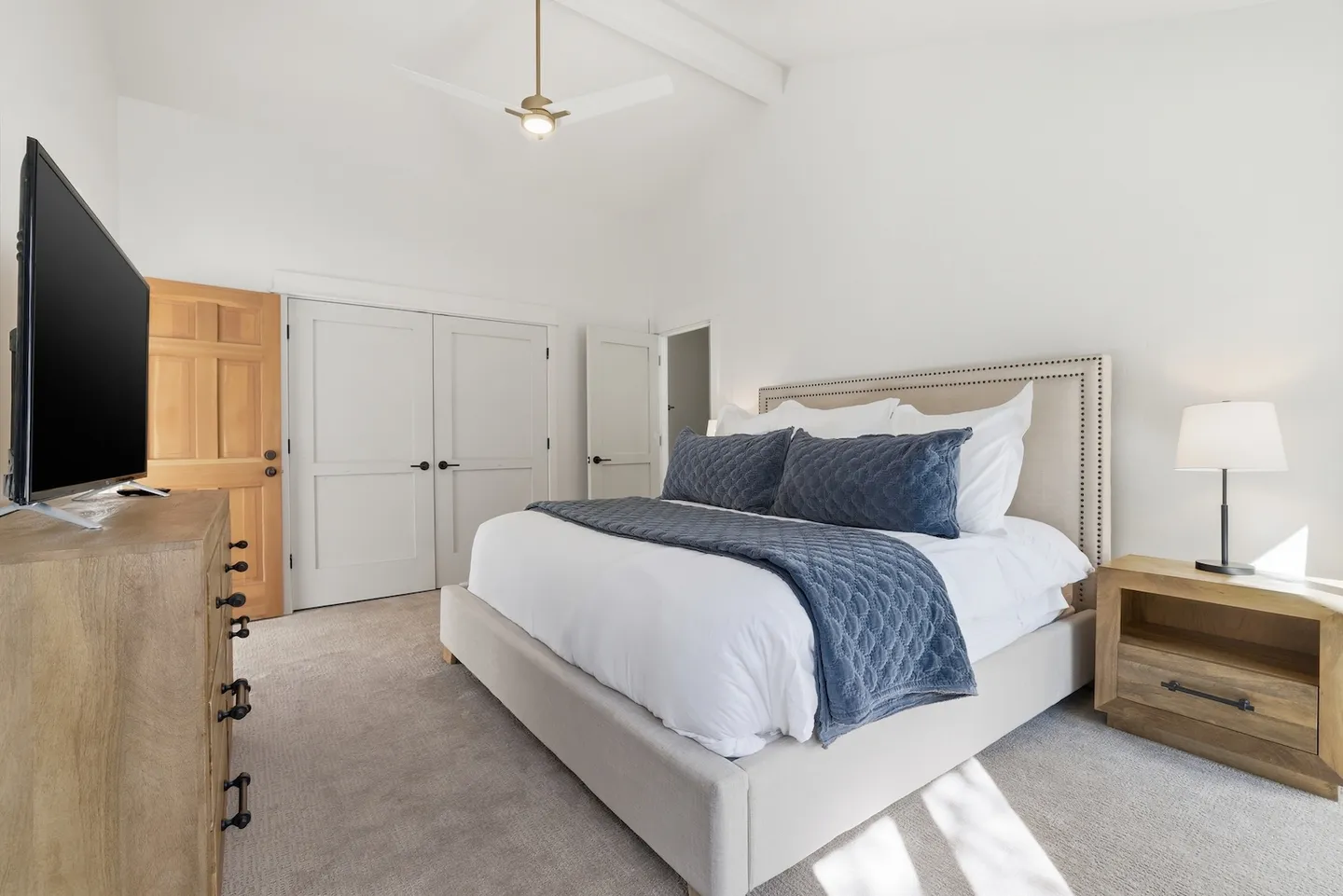 Bedroom with an upholstered bed, blue accent pillows, a ceiling fan, a dresser with a mounted television, and carpeted floors.