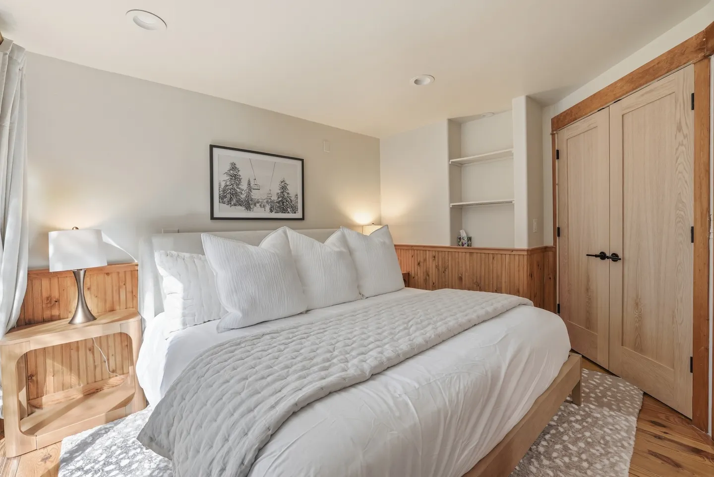 Bedroom with a large bed dressed in white linens, wood wainscoting along the walls, and a recessed shelving nook.