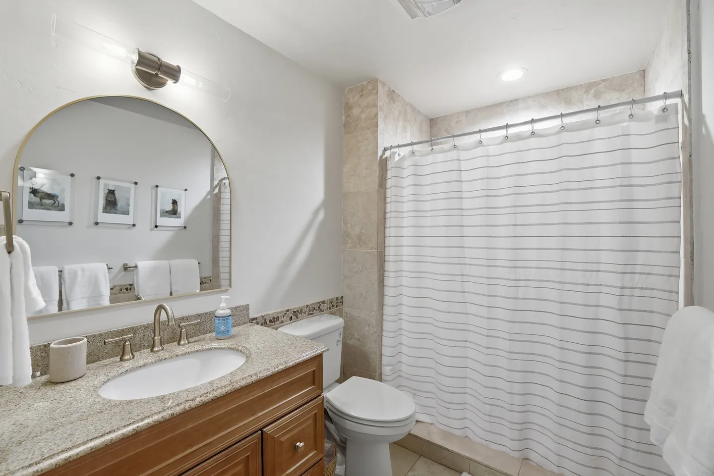 Bathroom with a wood vanity, arched mirror, toilet, and tiled shower area with a striped curtain.
