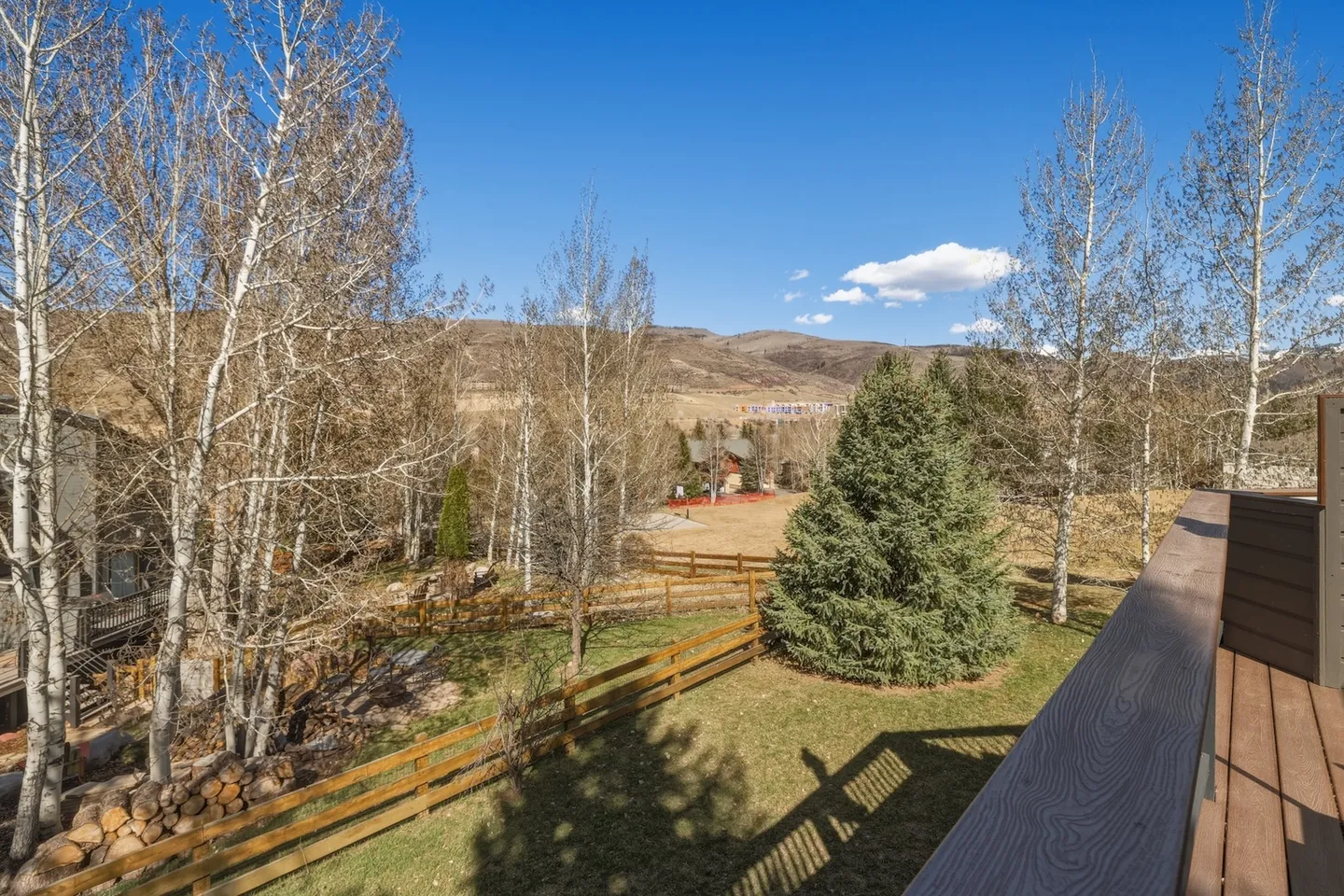 View from an elevated wooden deck looking out over a fenced yard with evergreen and deciduous trees against rolling hills and blue sky.