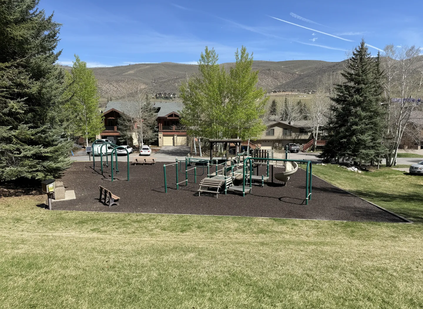 Outdoor community playground with climbing structures, slide, and swings surrounded by grass, trees, and residential buildings with mountains in the background.