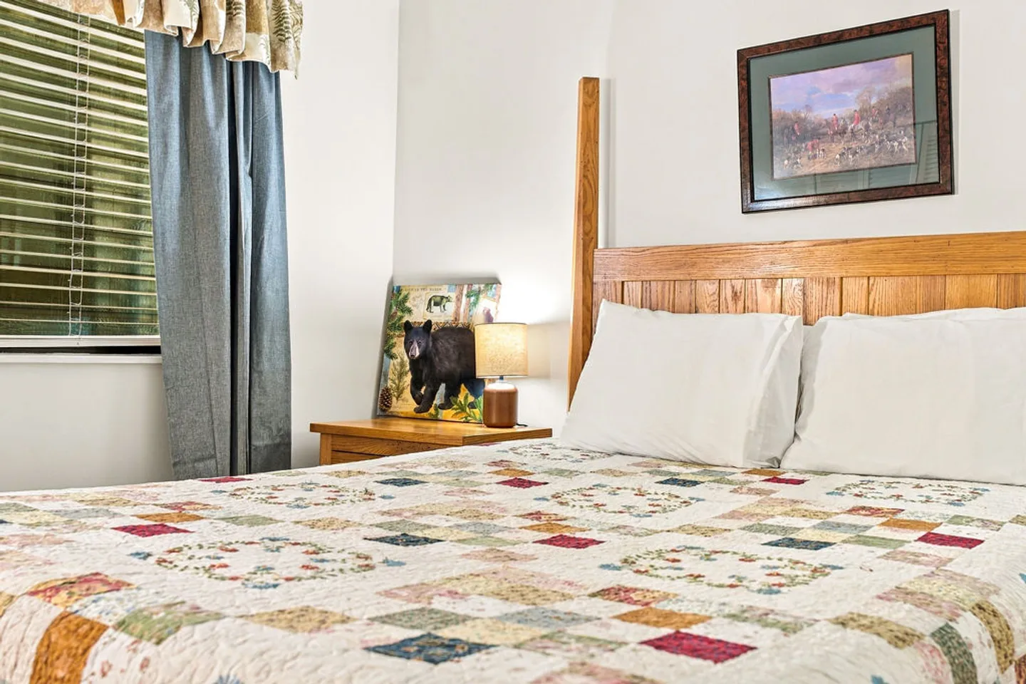 Bedroom with a wooden headboard, patchwork quilt, nightstand with a lamp, and window with blinds.