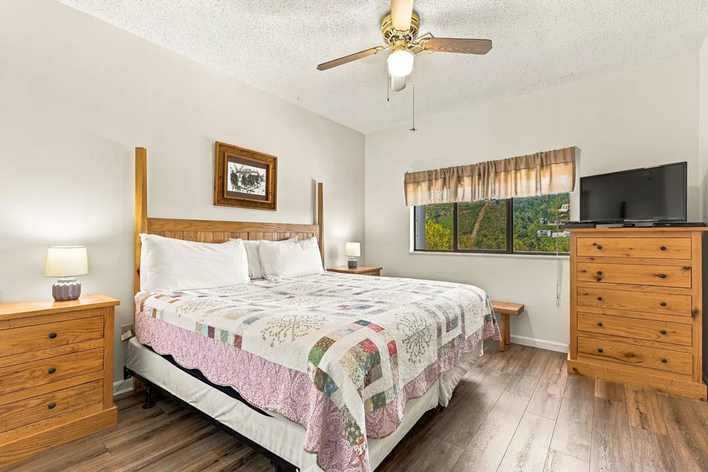 Bedroom featuring a wooden headboard, patchwork quilt, matching nightstands, a tall dresser with a TV, and a window with green outdoor views.