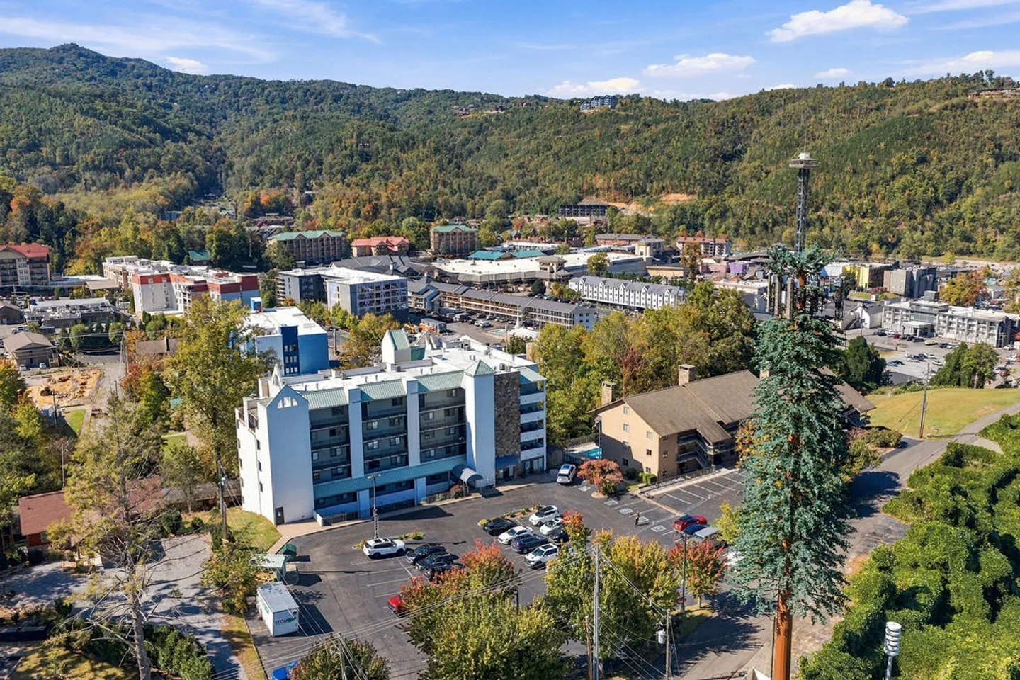 Aerial view of a multi-story resort or condo building surrounded by parking lots and a town against rolling forested mountains.