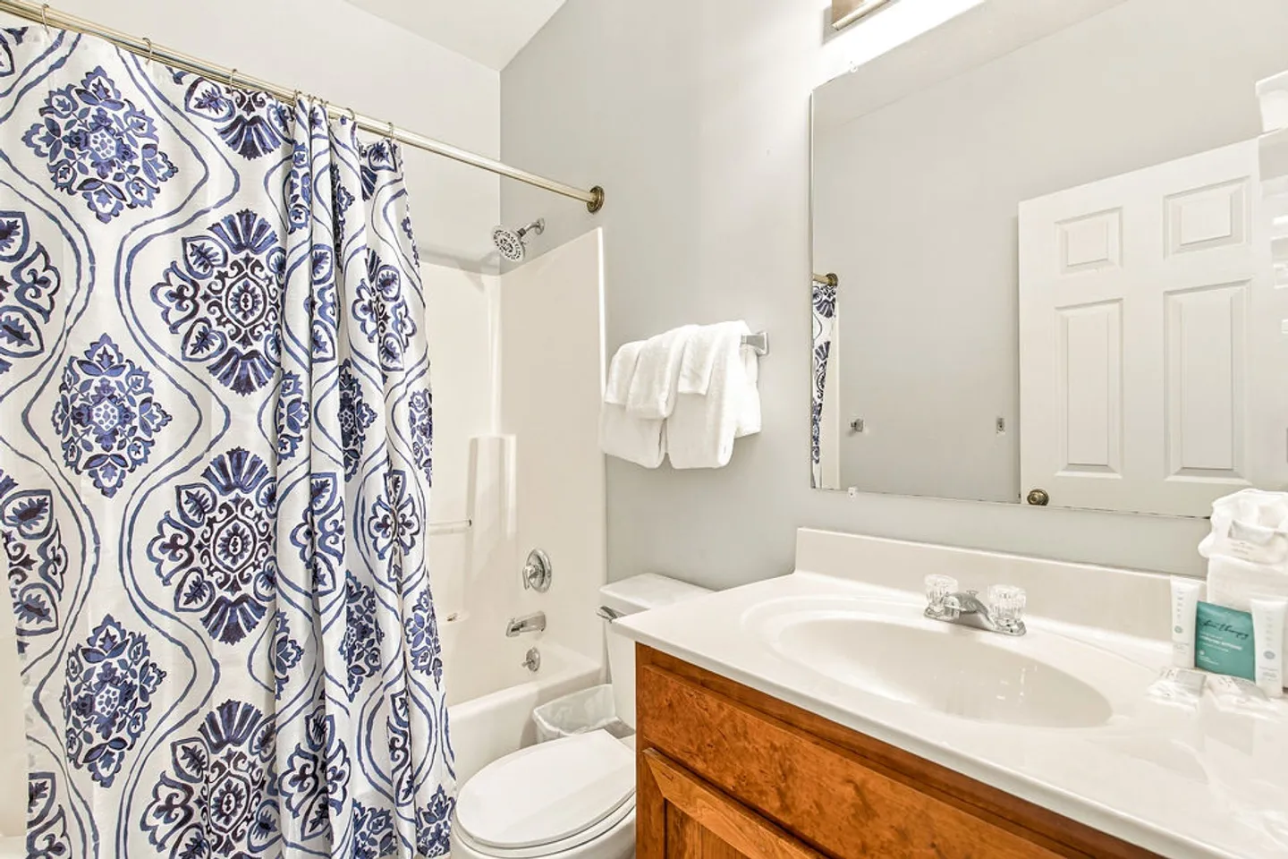 Bathroom showing a wooden vanity with a white countertop, toilet, and combination bathtub shower with a blue and white patterned curtain.
