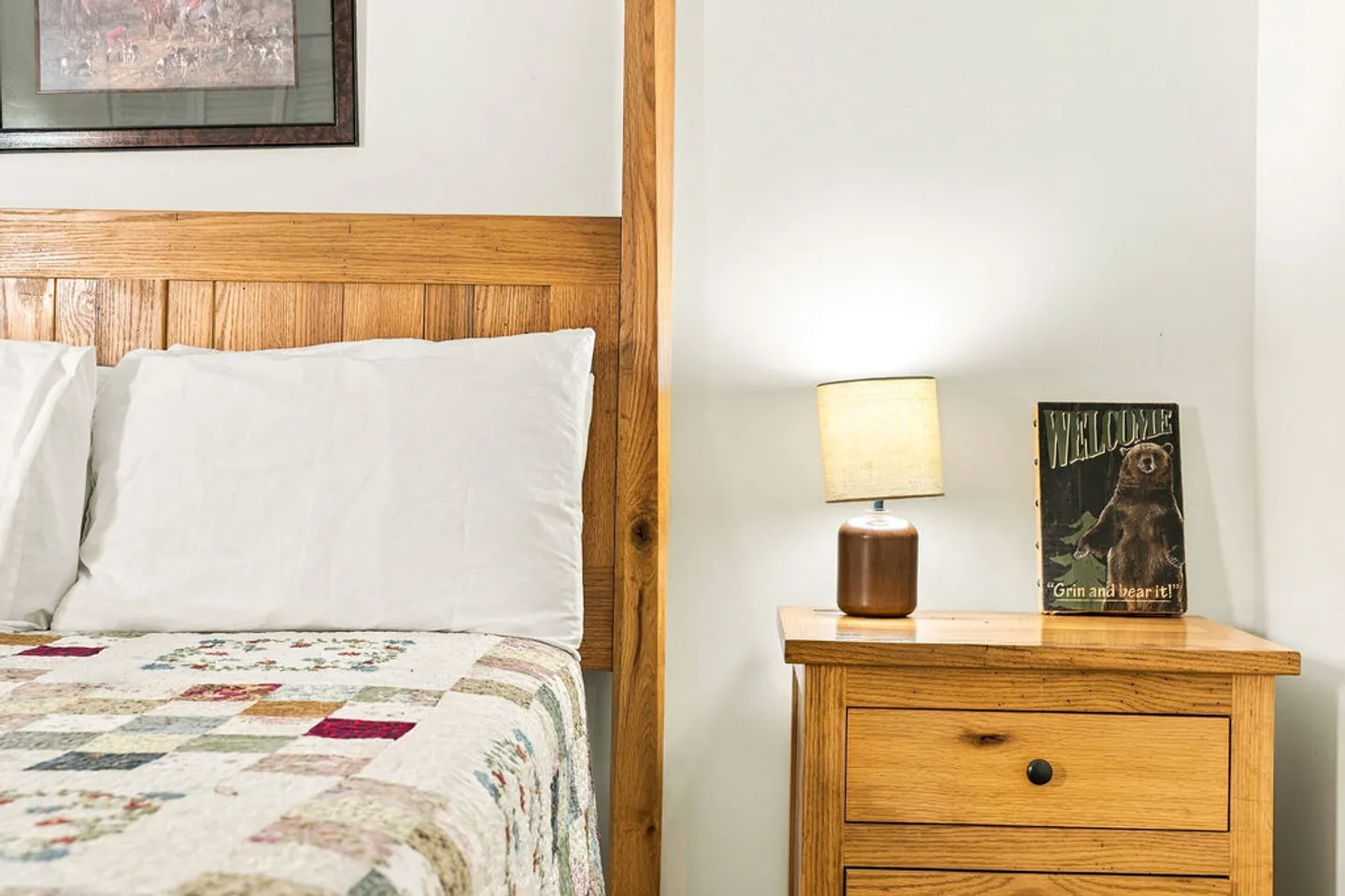 Close-up of a wooden bed frame with a patchwork quilt next to a wooden nightstand with an illuminated small lamp.