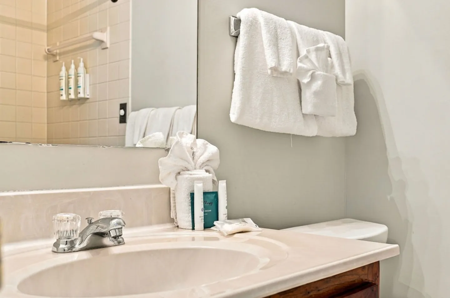 Bathroom vanity sink with chrome faucet, folded towels, toiletries, and mirror reflecting a tiled shower area.