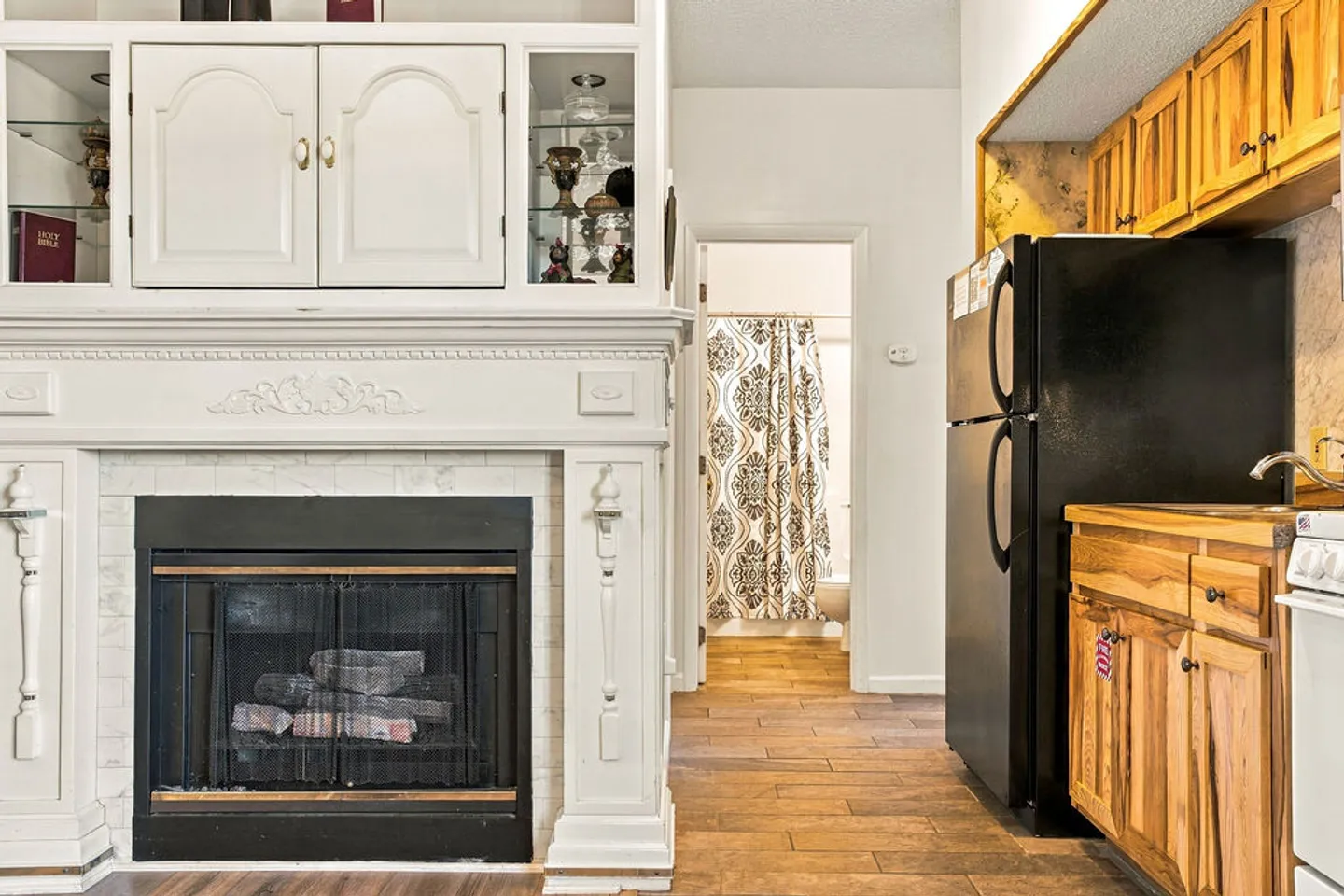 Living area showing a white fireplace mantel with built-in cabinetry next to a kitchen and doorway leading to a bathroom.