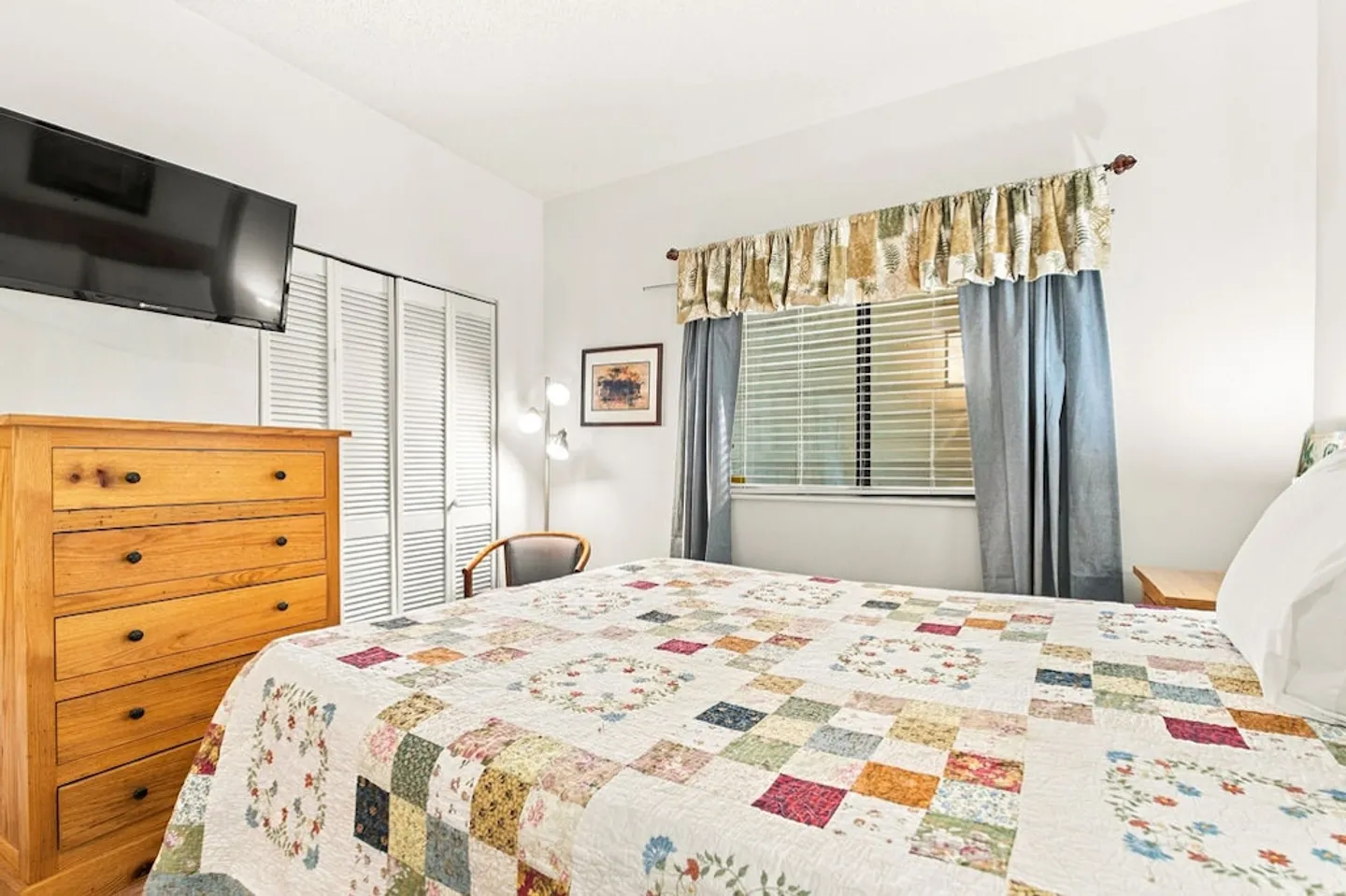 Bedroom with a patchwork quilt on the bed, wooden dresser, wall-mounted television, and a window with blinds.