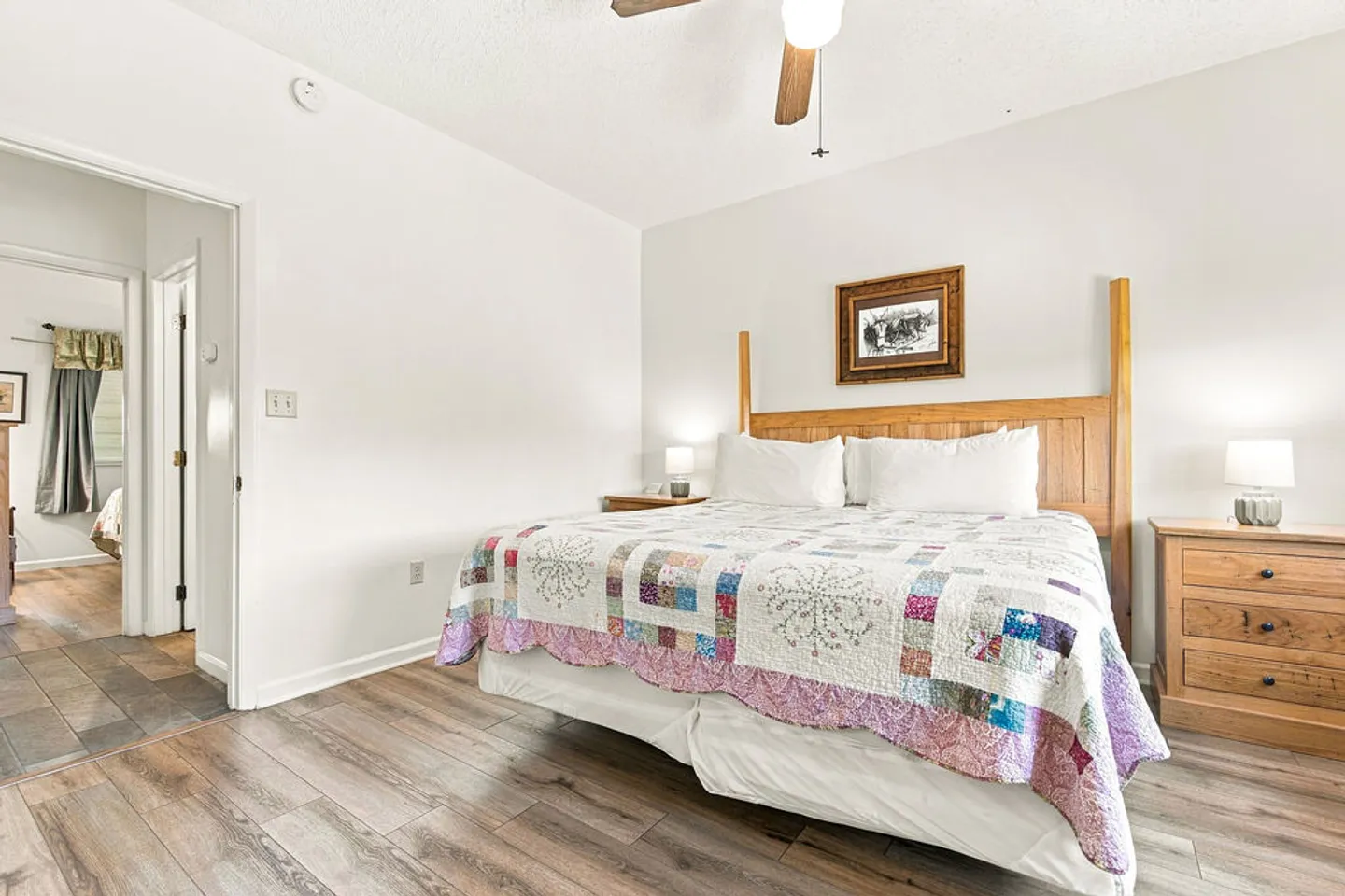 Bedroom with a bed dressed in a colorful quilt, a wooden headboard, two matching nightstands with lamps, and wood-look flooring.