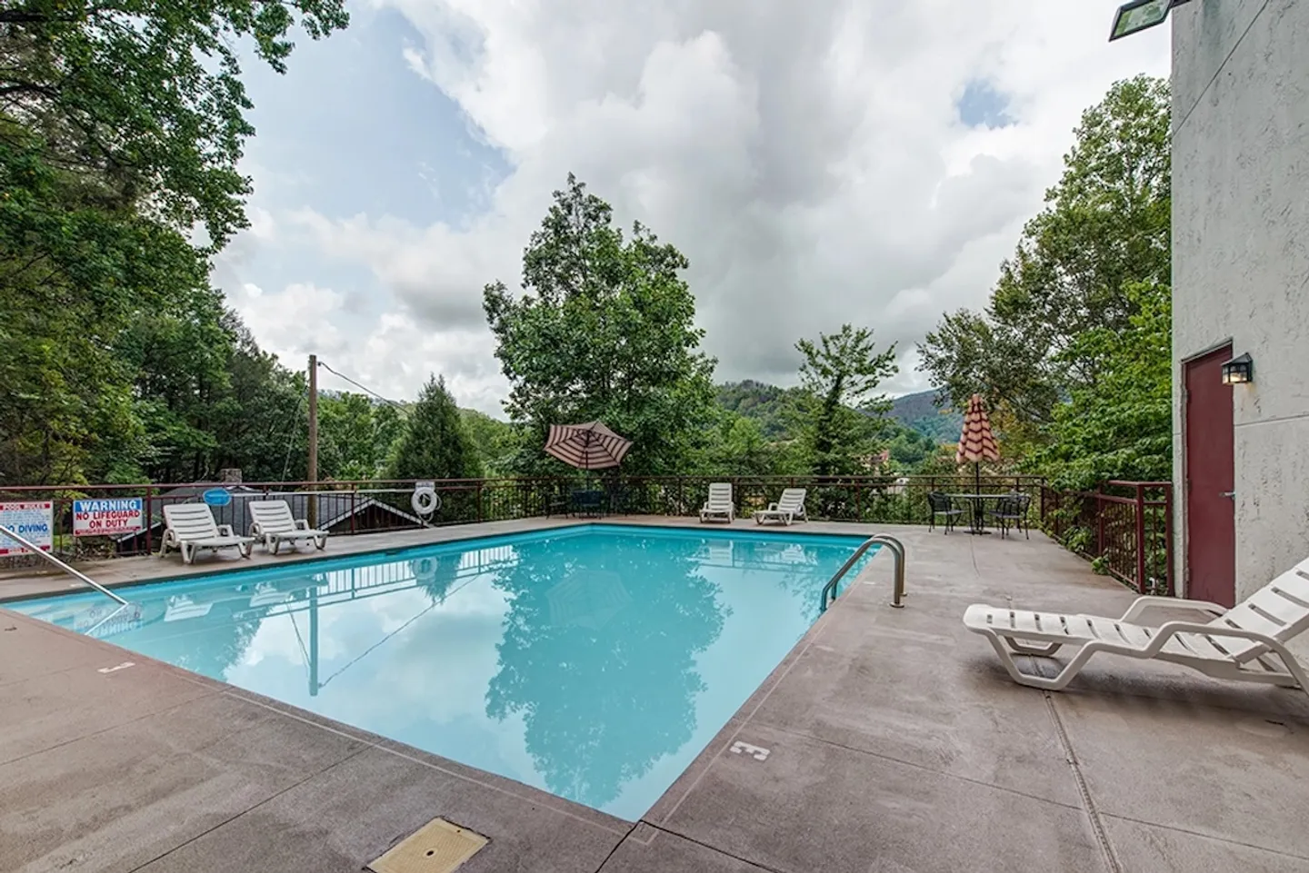 Outdoor swimming pool surrounded by a concrete deck with white lounge chairs, patio umbrellas, and tree-covered hills in the background.
