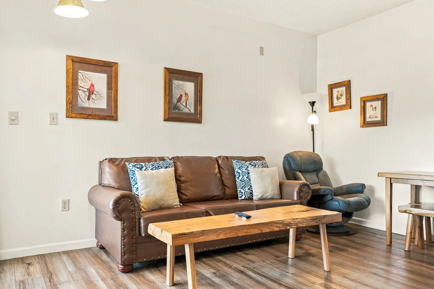 Living area featuring a brown leather sofa with accent pillows, a wooden coffee table, and an armchair against white walls.