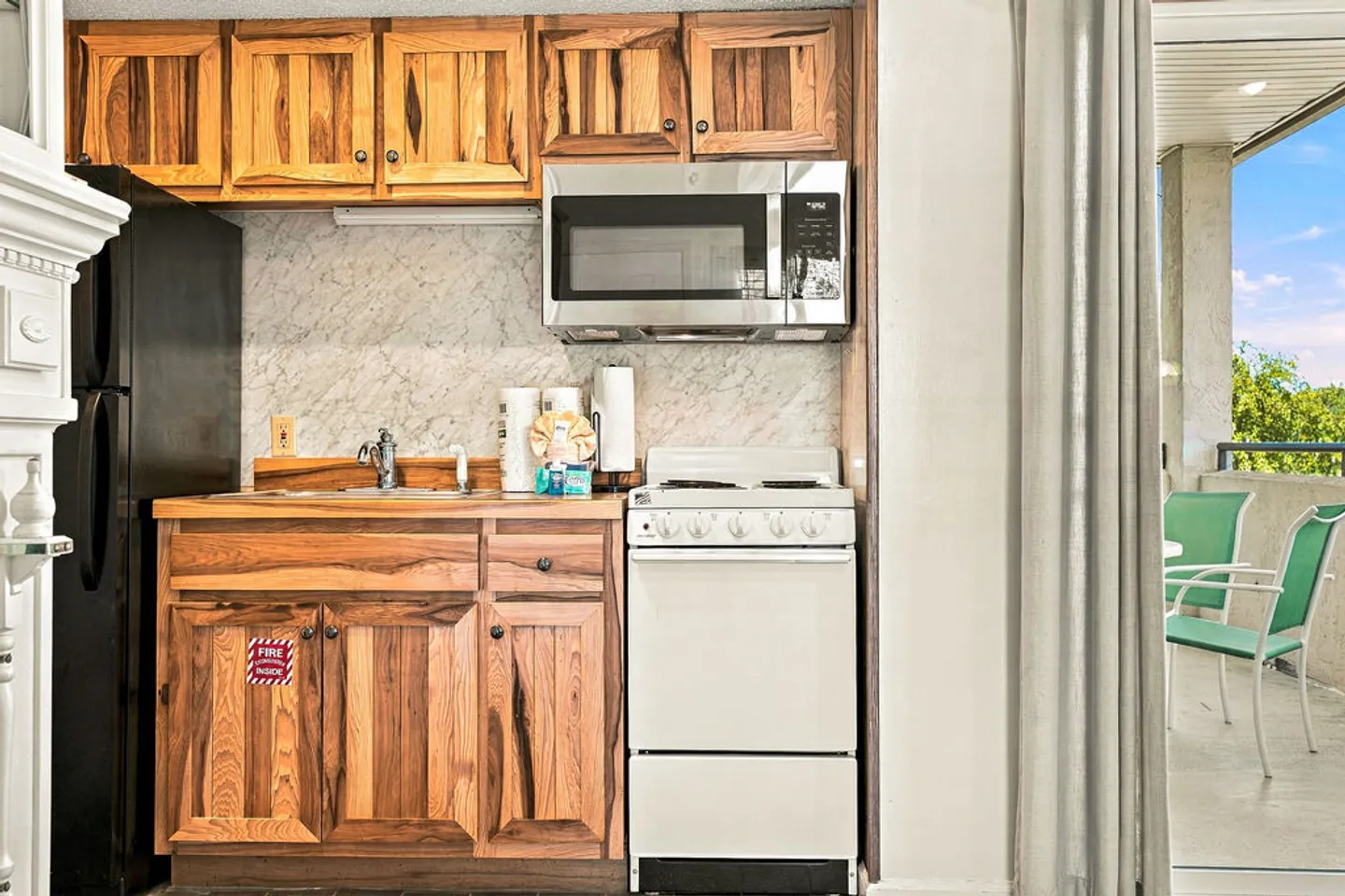Kitchenette with natural wood cabinets, white stove, microwave, and adjacent balcony with green chairs.