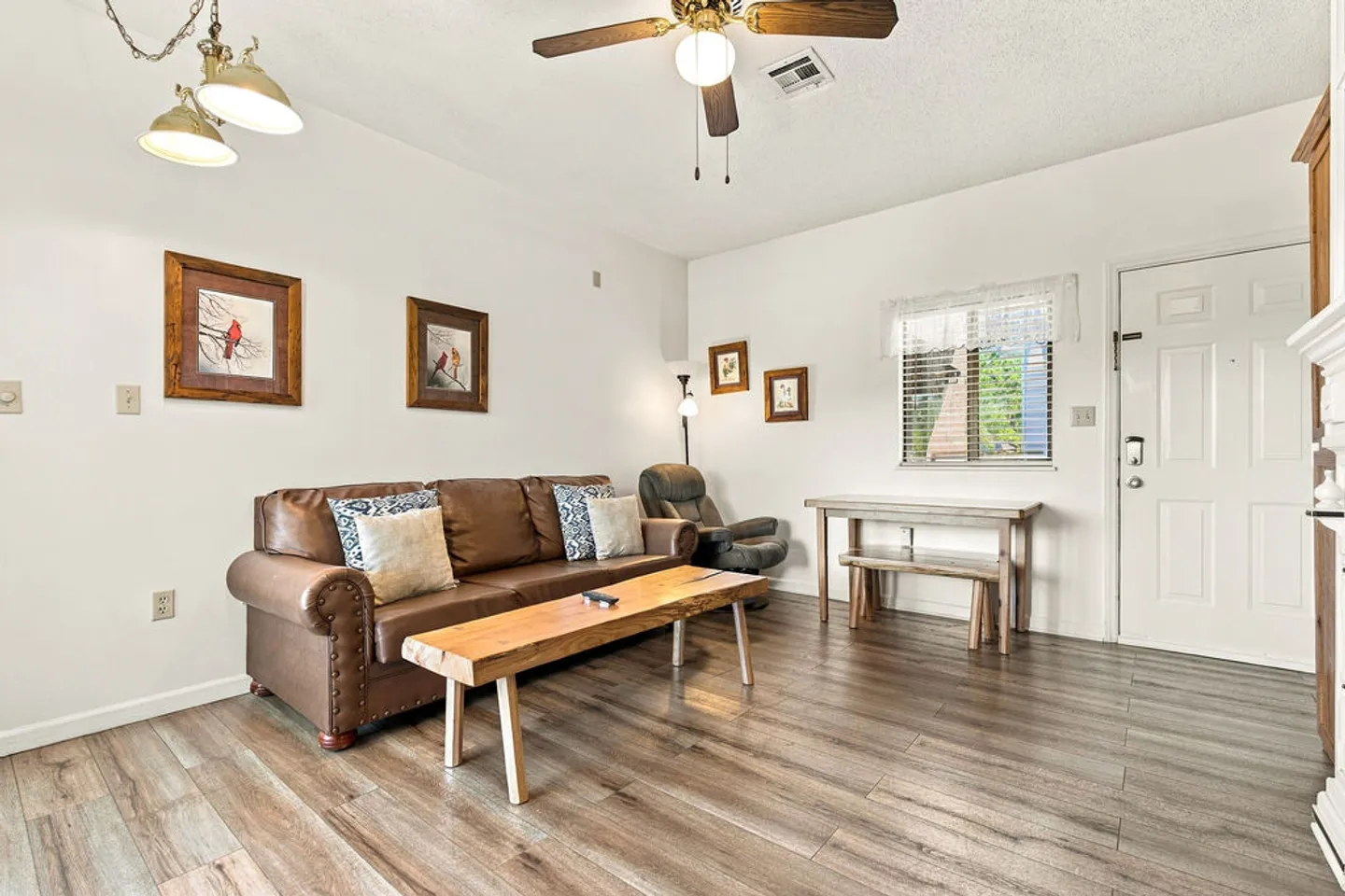 Living room featuring wood-look flooring, a brown leather sofa with a rustic coffee table, a ceiling fan, and an entrance door.