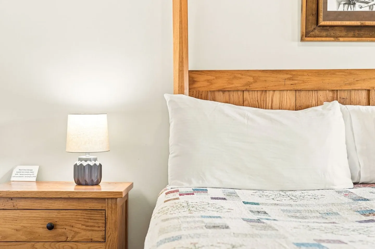 Close-up of a bed with a wooden headboard, patchwork quilt, and adjacent wooden nightstand with an illuminated small lamp.
