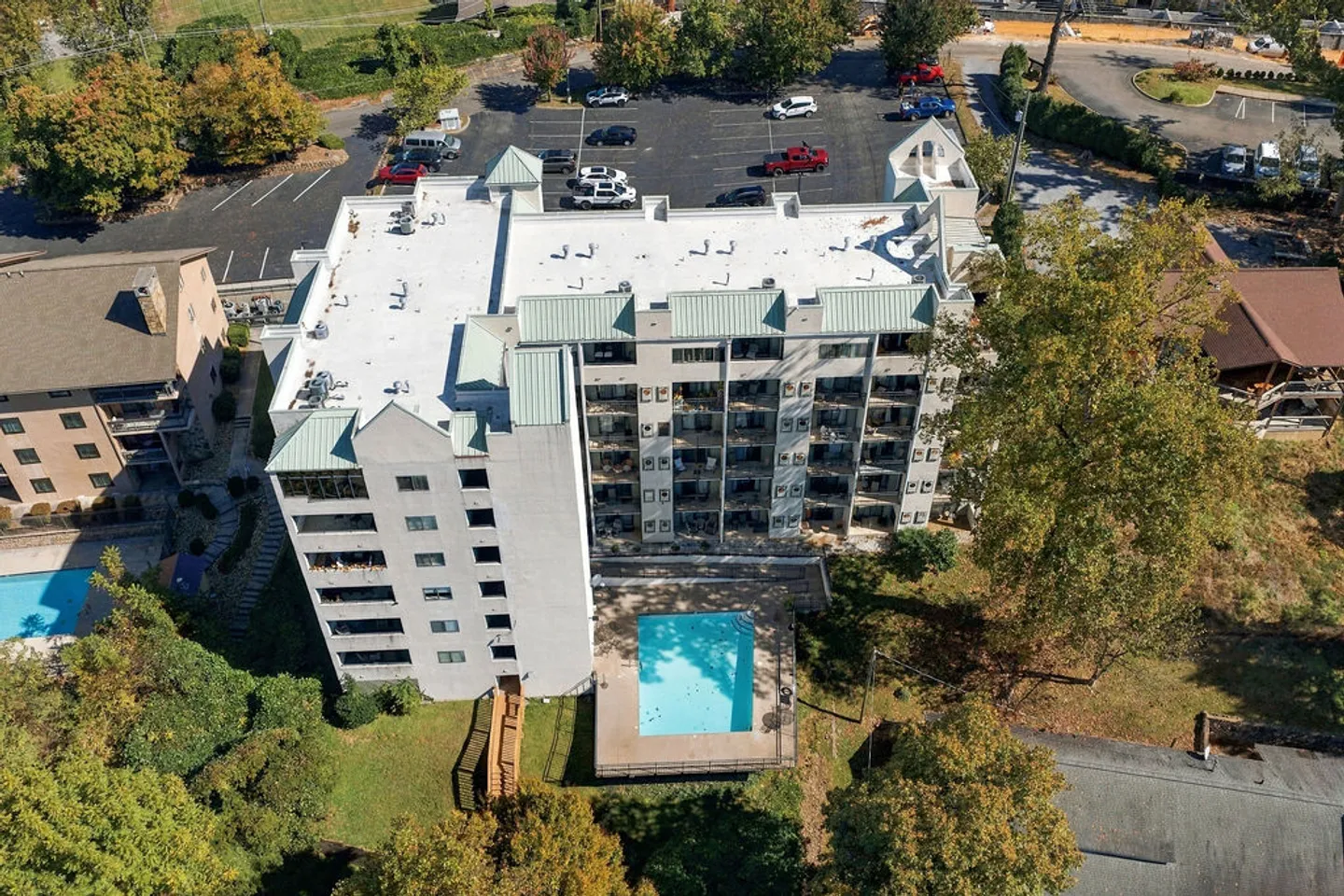 Aerial view of a multi-story condominium building featuring balconies and an outdoor swimming pool surrounded by trees and a parking lot.
