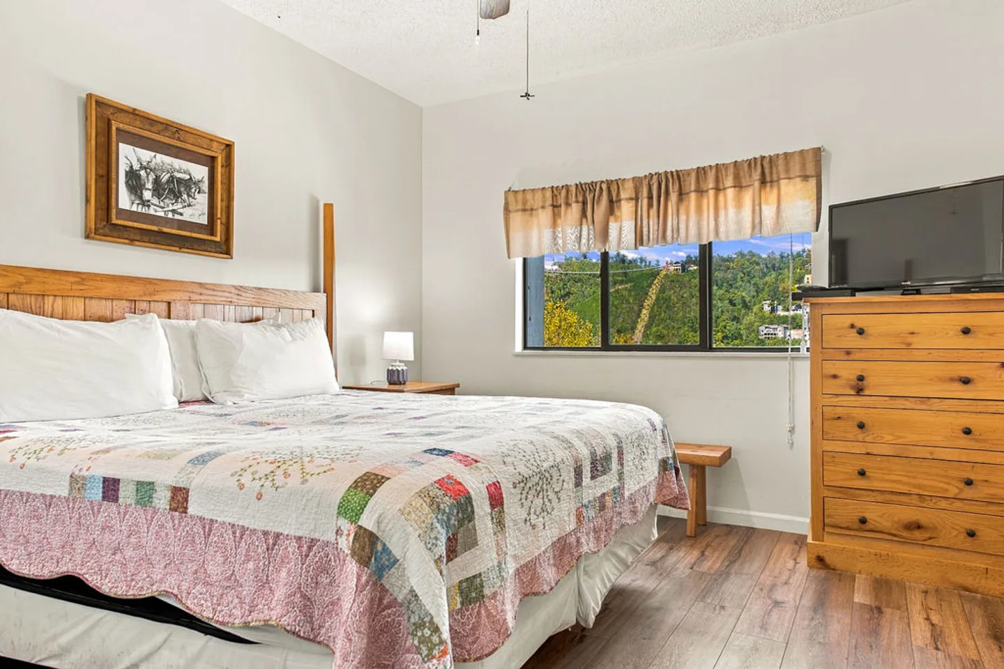 Bedroom with a wooden headboard, patchwork quilt on the bed, wooden dresser with a television, and window showing a mountain view.