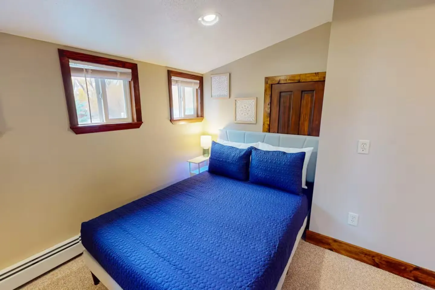 Bedroom with a bed made with a textured blue coverlet, carpeted floor, and two small windows with wood trim.