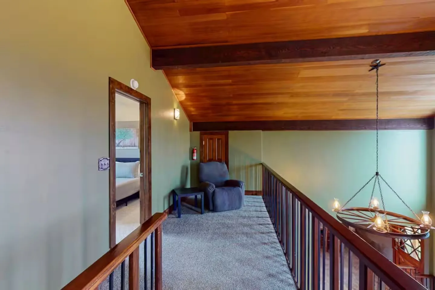 Upstairs loft hallway with carpet, wood railing, wood-paneled vaulted ceiling, and an armchair next to a bedroom doorway.