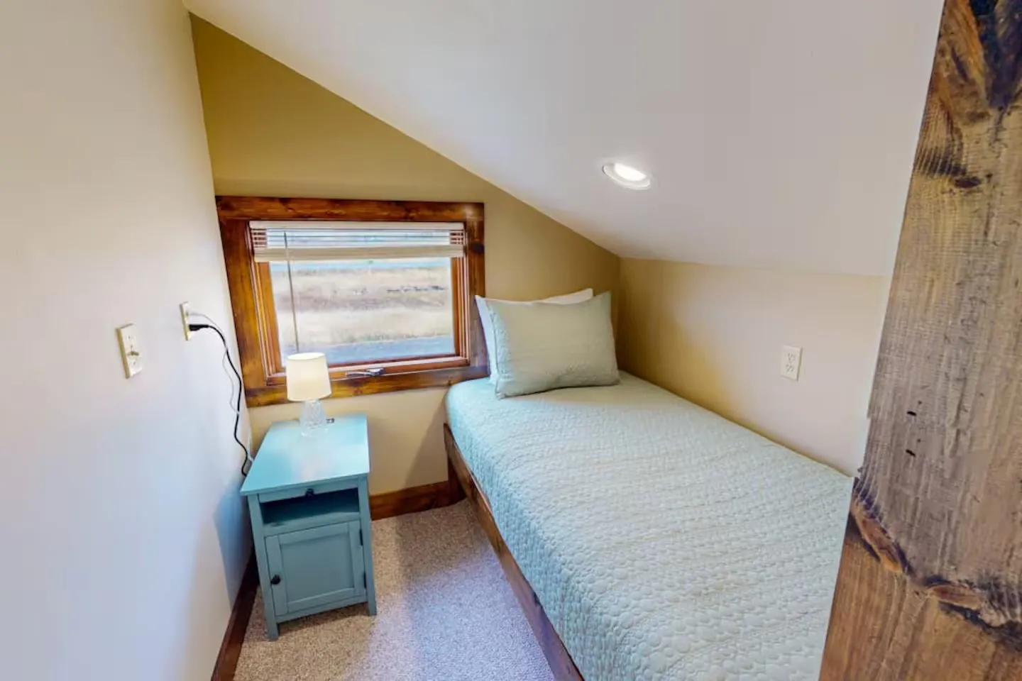 A cozy bedroom alcove beneath a sloped ceiling with a single bed, small turquoise nightstand, and a wood-trimmed window.