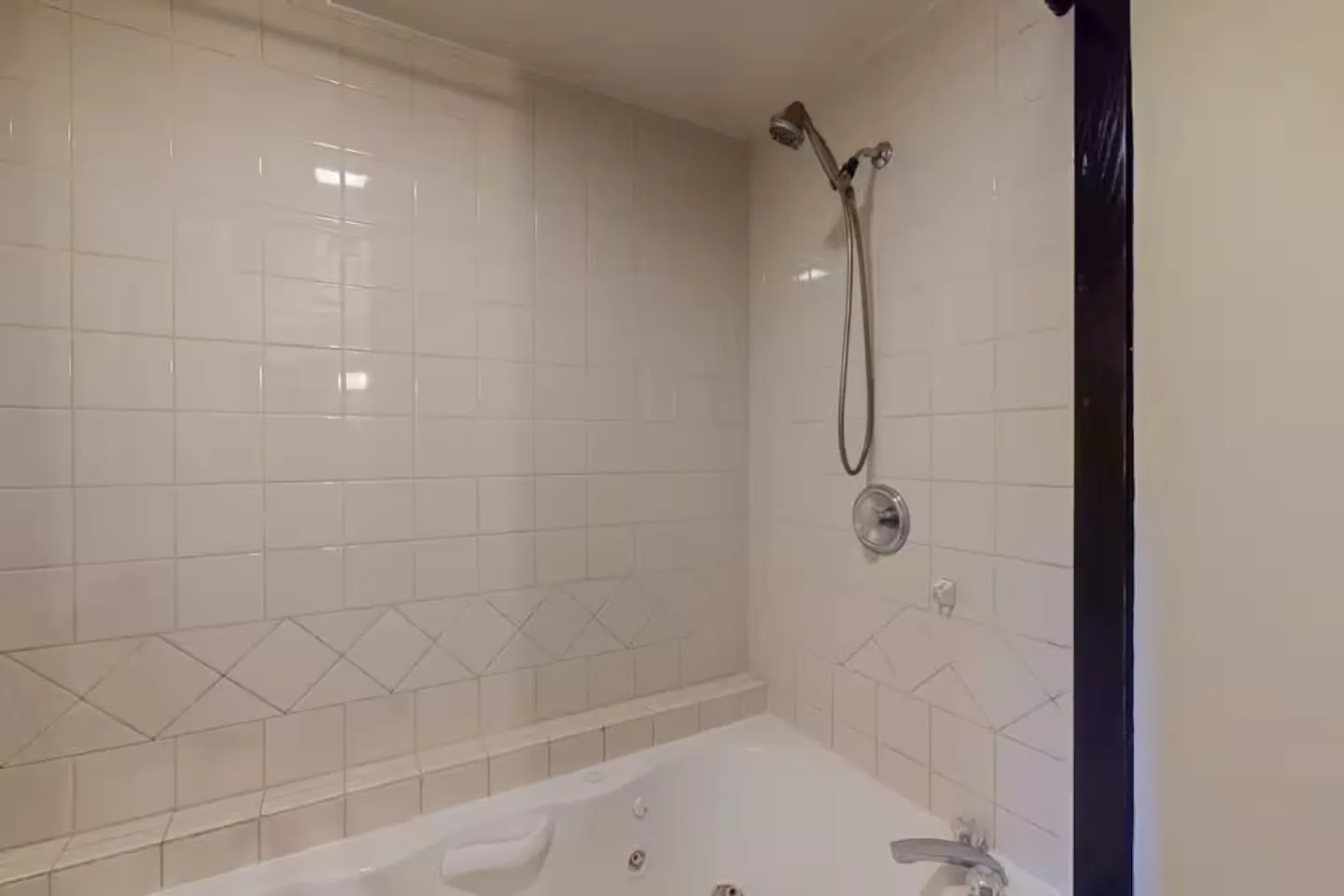 A close-up view of a bathroom jetted tub with tiled walls and a handheld showerhead attachment.