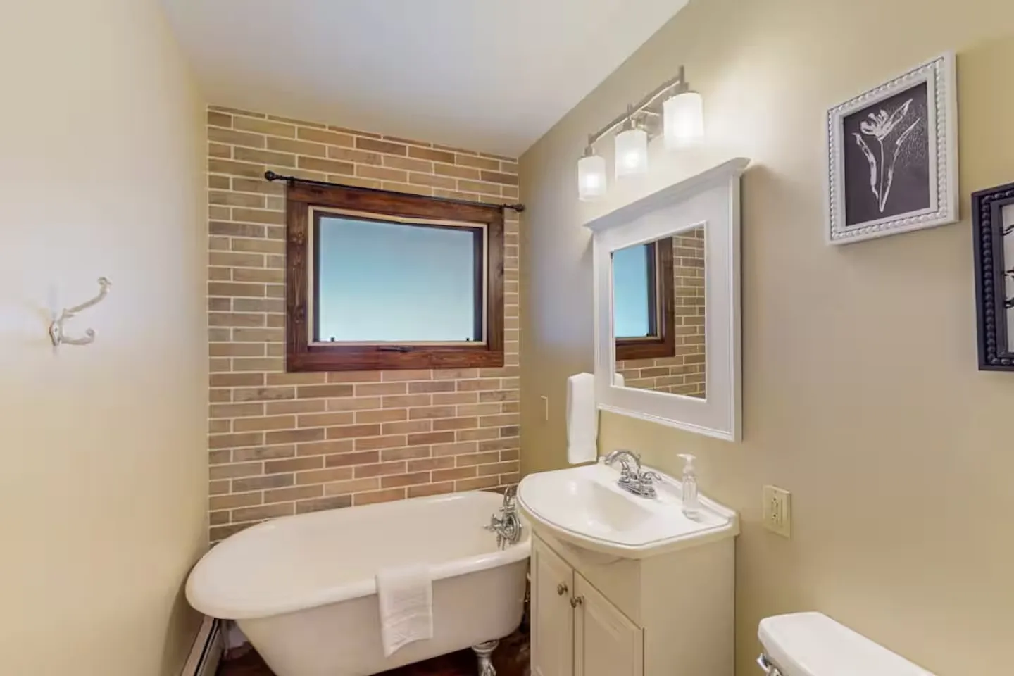Bathroom featuring a freestanding clawfoot bathtub, vanity sink with mirror, and brick-look accent wall with a window.