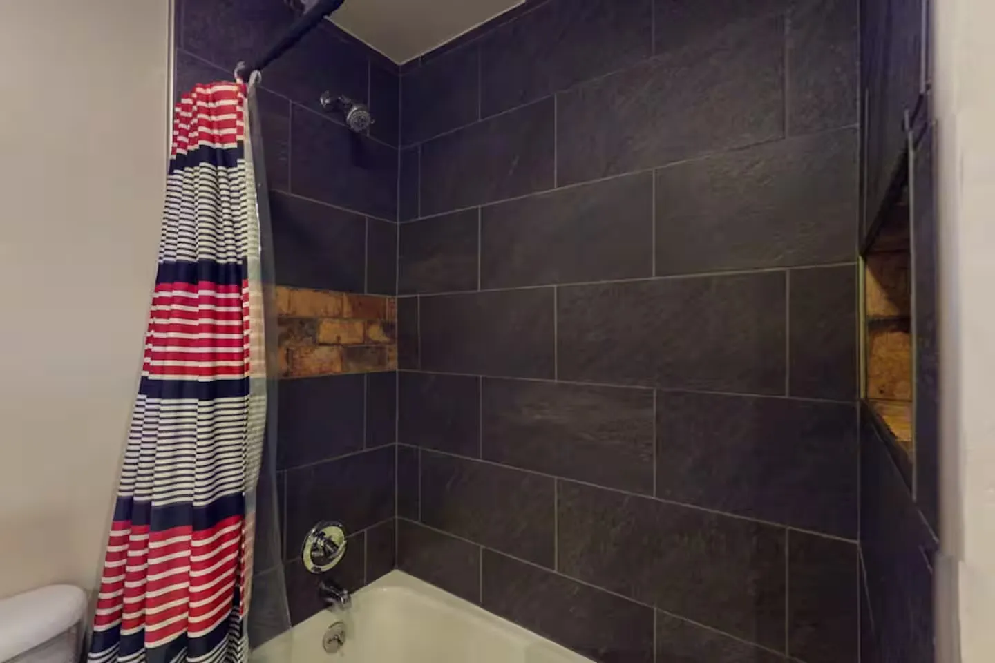 Bathroom shower and tub combination featuring dark slate-style tile, an exposed brick accent patch, and a striped shower curtain.
