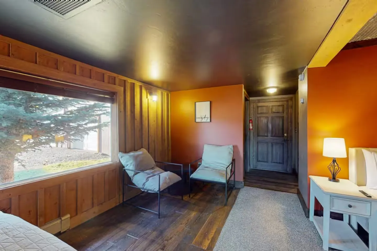 Seating area in a rustic bedroom featuring a large window, dark ceiling, wood-paneled accents, and warm orange walls.