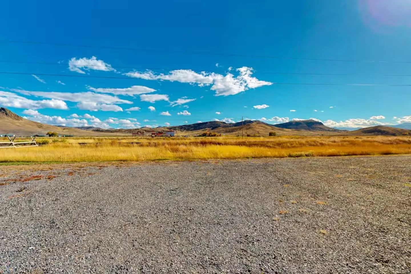 Open gravel lot overlooking a golden field with distant hills under a bright blue sky with clouds.