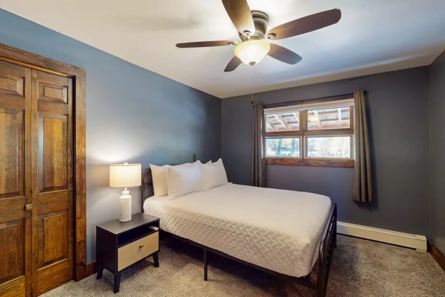 Bedroom with blue walls, carpeted floor, bed with white bedding, wooden bifold closet doors, and a ceiling fan.