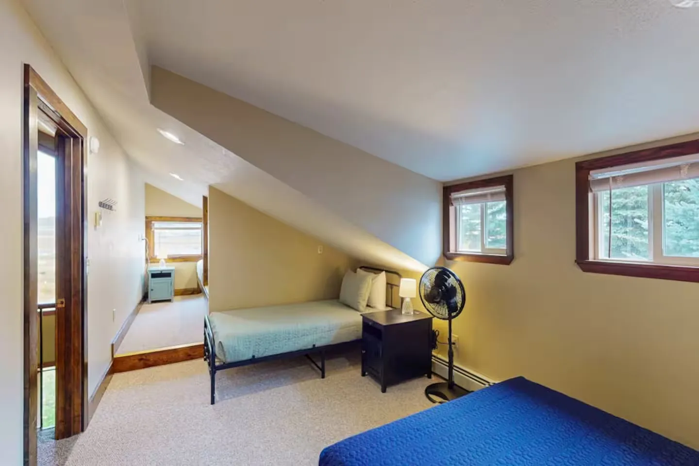 Upstairs bedroom with sloped ceilings, carpeted flooring, twin-style beds, a nightstand with a lamp, and windows with wooden trim.
