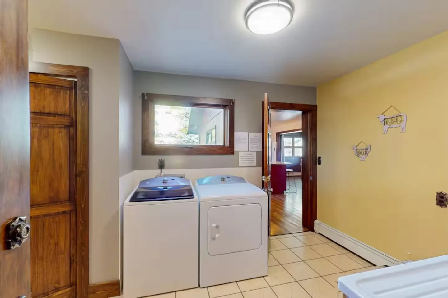 A laundry room with a white washer and dryer, light tile flooring, a window, and an open doorway leading to another room.