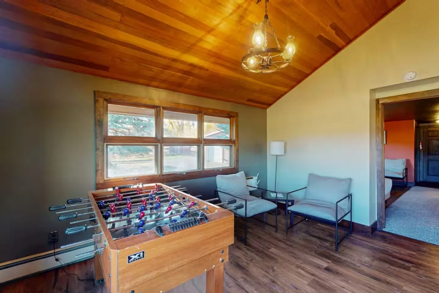 Game area with a foosball table, two armchairs, wood floors, and a wood-paneled angled ceiling with a wagon wheel light fixture.