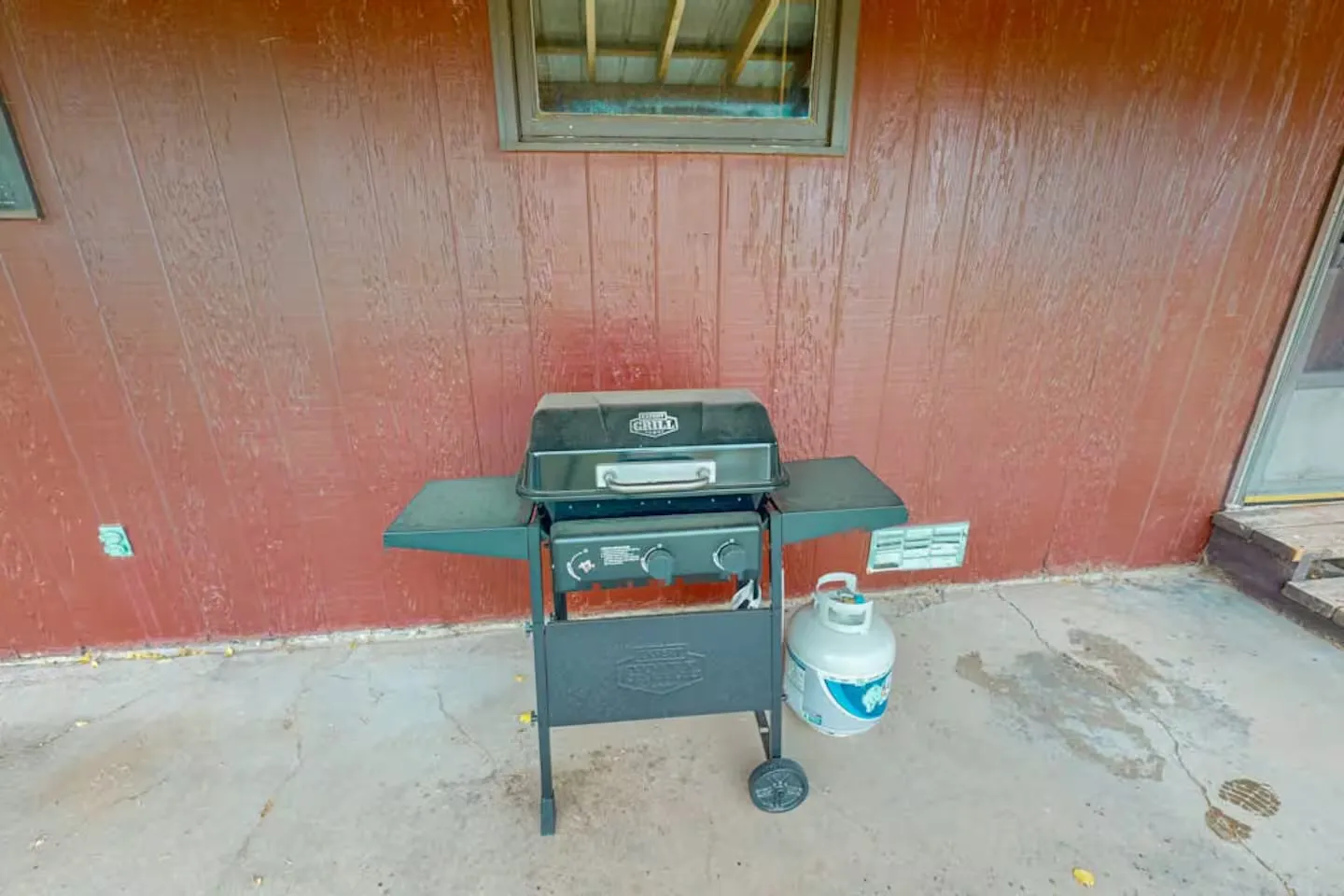 A black gas grill with a propane tank sits on a concrete patio in front of a red wooden exterior wall.