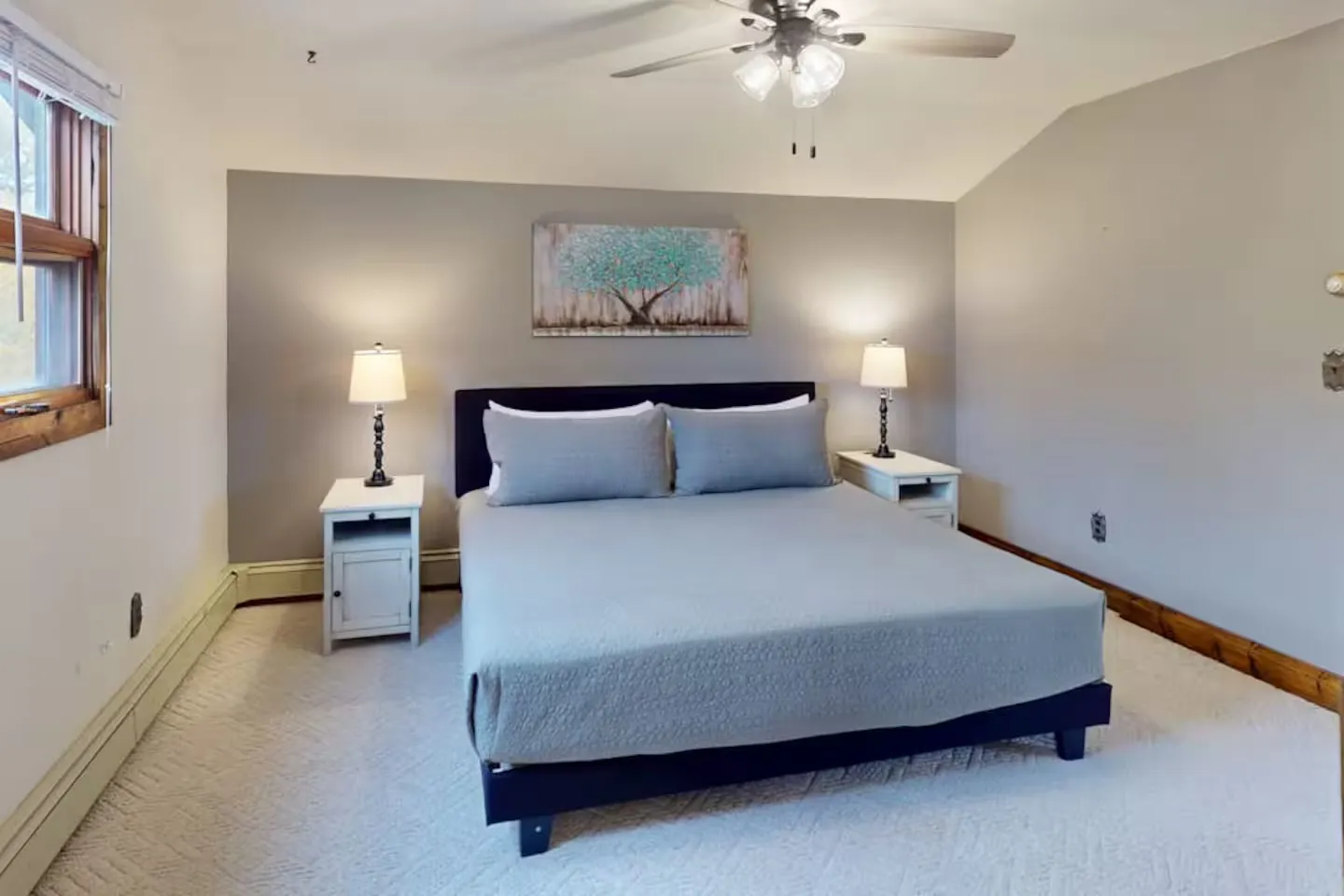 Bedroom with a bed flanked by nightstands and lamps, carpeted floor, ceiling fan, and painted tree artwork on the wall.