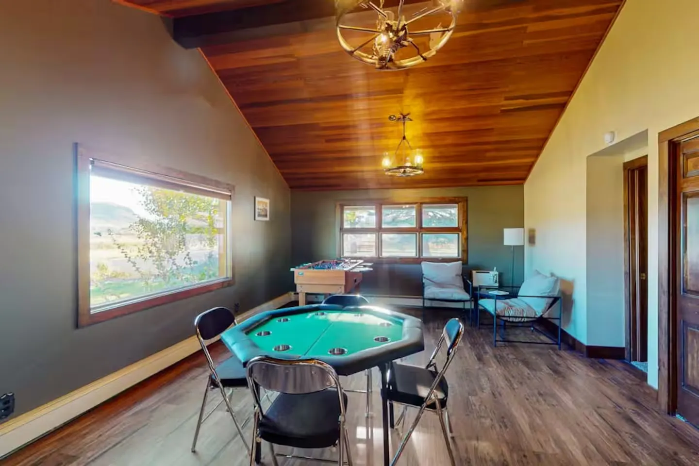 Game room featuring a green octagonal poker table with chairs, a foosball table, wood plank ceiling, and windows looking outside.