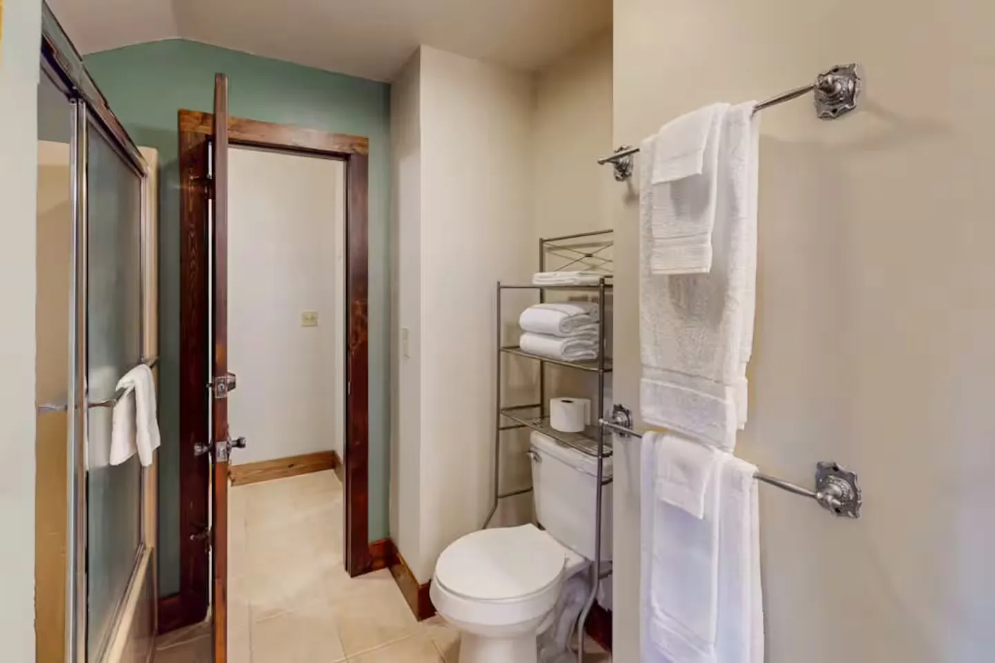 Bathroom with a toilet, over-the-toilet metal shelf with towels, sliding glass shower doors, and an open doorway.