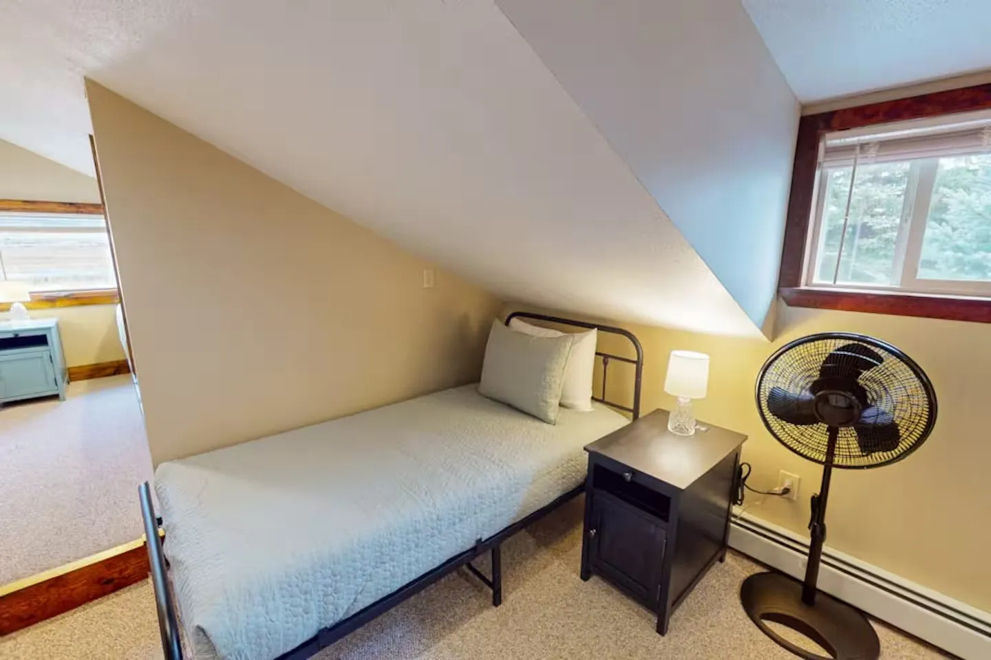 A twin bed tucked under a sharply sloped ceiling with a small nightstand, lamp, and floor fan nearby.