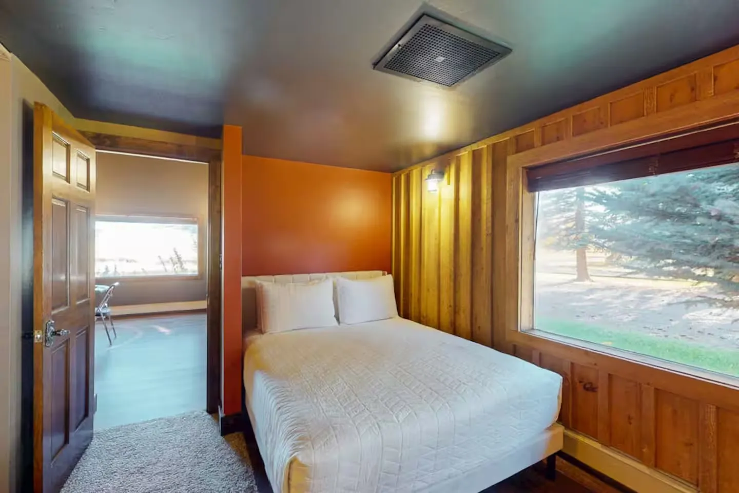 Bedroom with wood-paneled wall, orange accent wall, and a bed next to a large picture window.