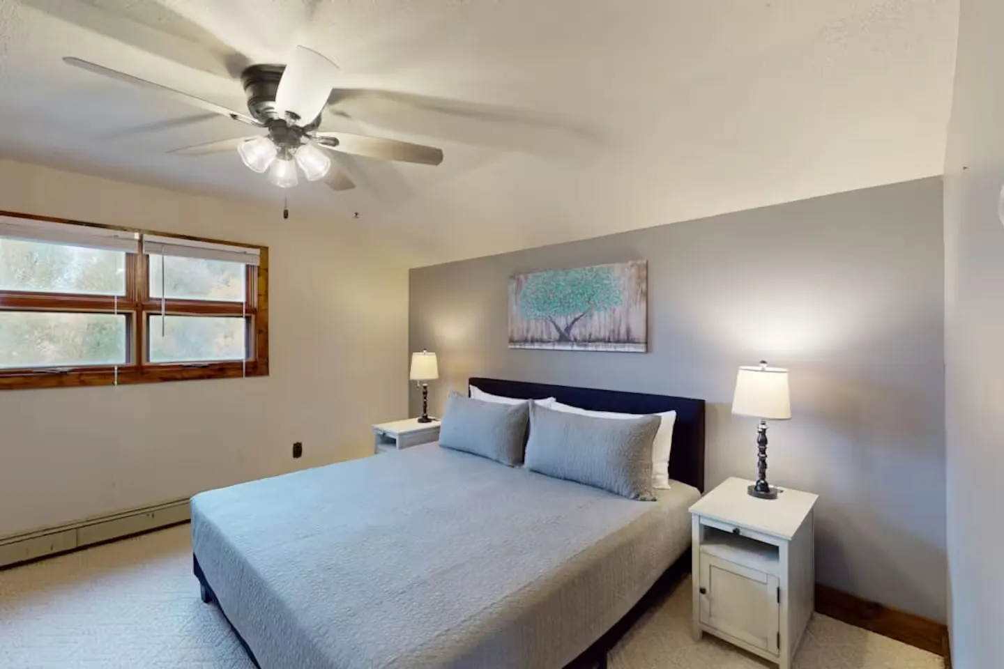 Bedroom featuring a made bed with gray bedding, matching nightstands with lamps, a ceiling fan, and a window.