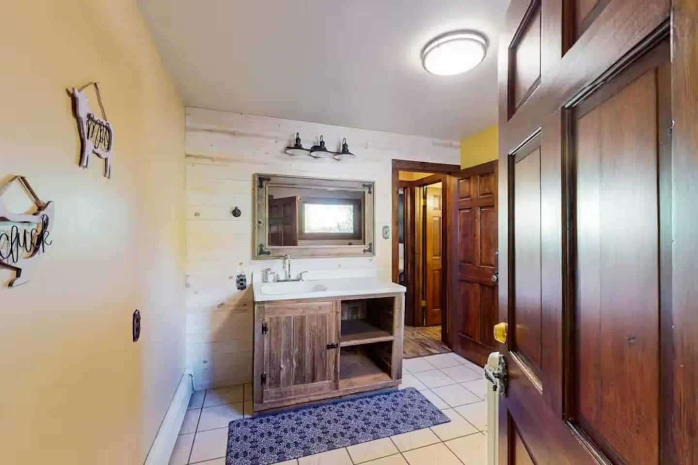 Bathroom with a rustic wooden vanity, a framed mirror, shiplap accent wall, tiled floor, and an open wooden door.