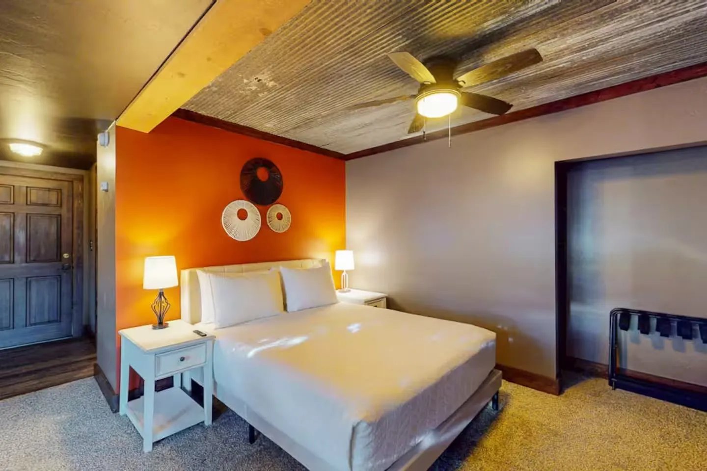 Bedroom featuring a bed with white linens, a bright orange accent wall, and a corrugated metal ceiling with a ceiling fan.