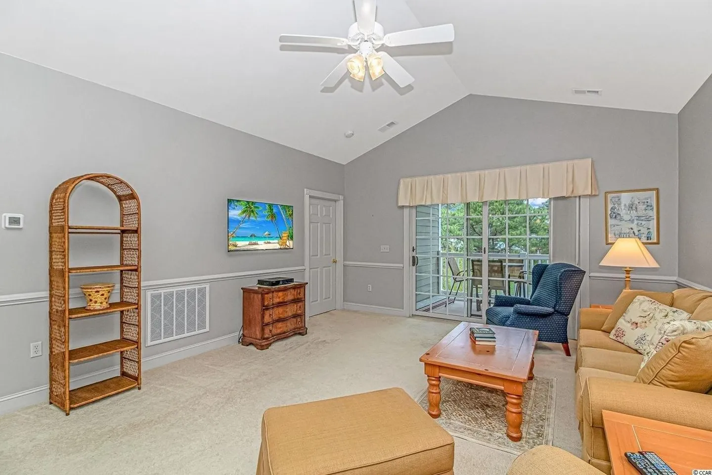 Living room with vaulted ceiling, carpeted floor, sofa, armchair, coffee table, wall-mounted television, and sliding glass door leading to a screened porch.