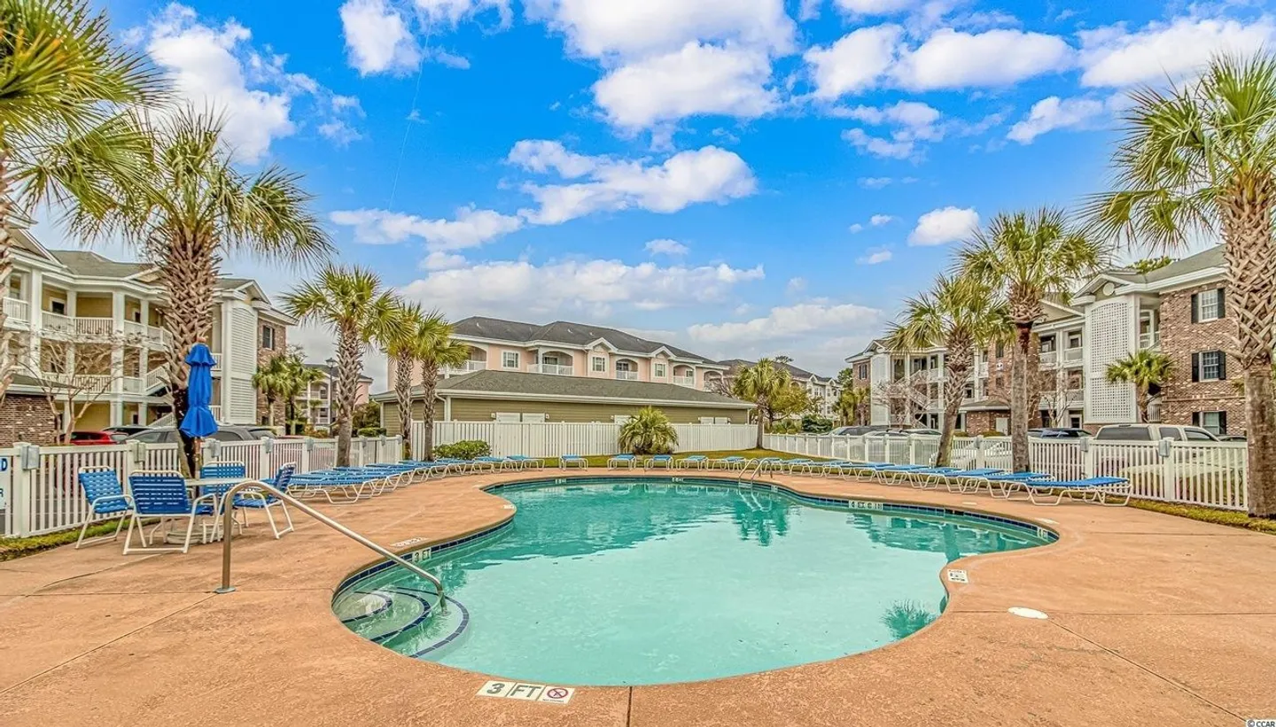 Outdoor curved swimming pool surrounded by lounge chairs, palm trees, and residential apartment buildings under a partly cloudy sky.