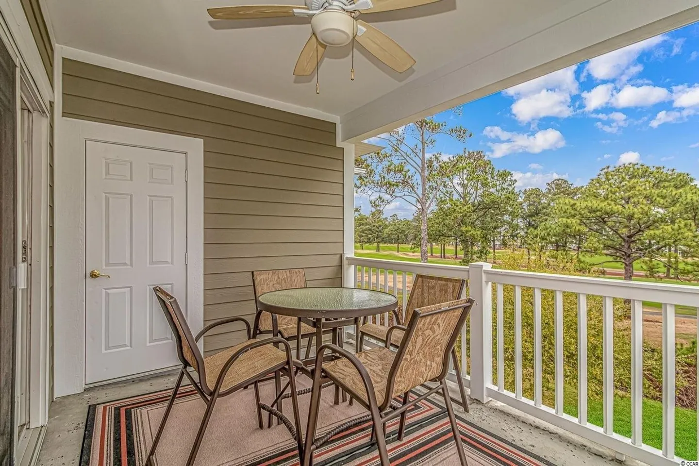 Covered balcony featuring a round outdoor table, chairs, an area rug, a ceiling fan, and views of a green lawn and trees.